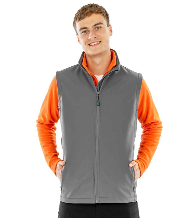 A young person faces forward with hands in the pockets of a gray Result Recycled Printable Soft Shell Bodywarmer by Result Recycled, layered over a bright orange long-sleeve shirt against a plain white background.