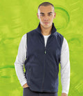A person stands facing forward, wearing the Result Recycled Polarthermic Fleece Bodywarmer in navy over a white long-sleeve shirt with hands in fleece-lined pockets. The bright green background features abstract swirls. Brand: Result Recycled.