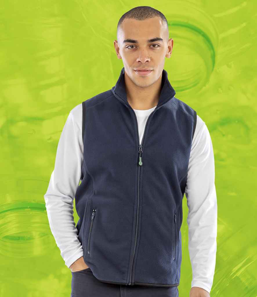 A person stands facing forward, wearing the Result Recycled Polarthermic Fleece Bodywarmer in navy over a white long-sleeve shirt with hands in fleece-lined pockets. The bright green background features abstract swirls. Brand: Result Recycled.
