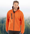 A man wearing the Result Recycled Three Layer Printable Hooded Soft Shell Jacket by Result Recycled, featuring an orange waterproof design with a black zipper and zippered chest pocket, stands before a misty forest, uniting style and function.