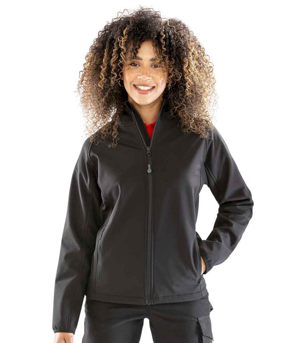 A woman with curly hair stands smiling against a white background, wearing the Result Recycled Ladies Three Layer Printable Hooded Soft Shell Jacket in black by Result Recycled, paired with black pants.