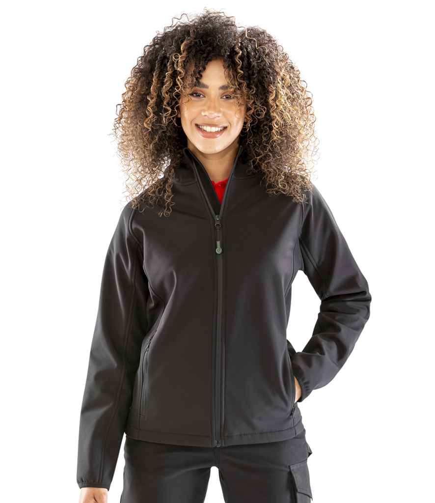 A woman with curly hair stands smiling against a white background, wearing the Result Recycled Ladies Three Layer Printable Hooded Soft Shell Jacket in black by Result Recycled, paired with black pants.