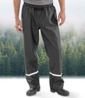A person wearing Result Recycled Prism PU Waterproof Trousers by Result Recycled and black boots stands before a misty evergreen forest, the trousers featuring reflective bands near the ankles.