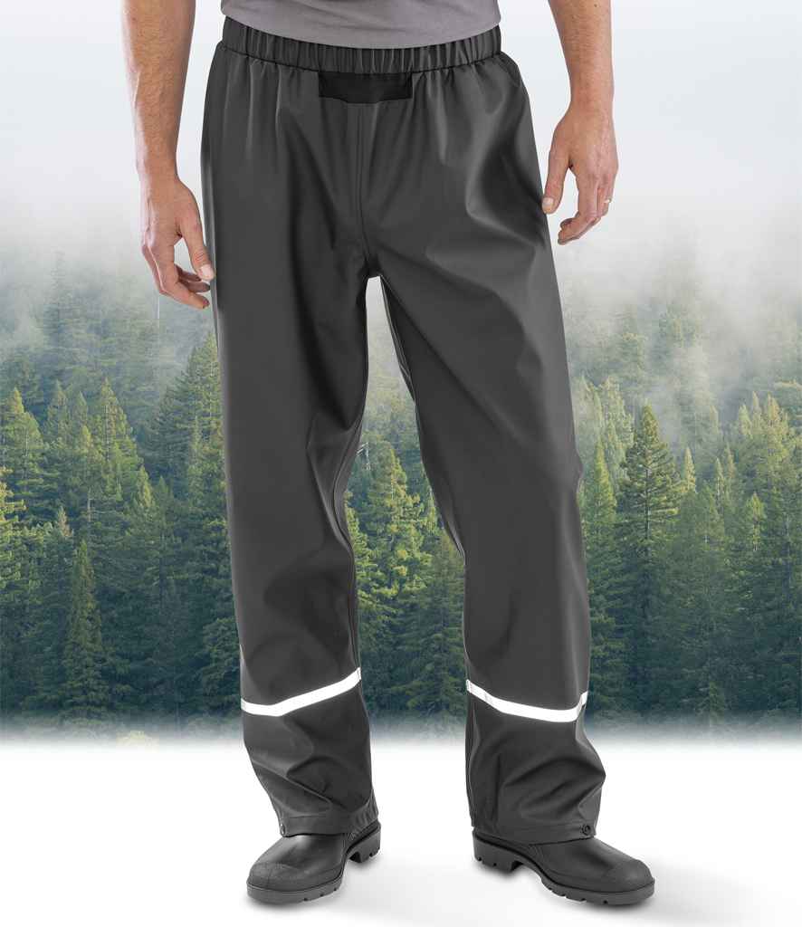 A person wearing Result Recycled Prism PU Waterproof Trousers by Result Recycled and black boots stands before a misty evergreen forest, the trousers featuring reflective bands near the ankles.