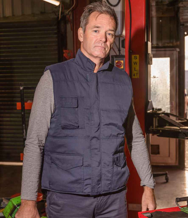 A middle-aged man with short brown hair wears the Result Promo Bodywarmer by Result over a long-sleeve gray shirt, standing indoors in a workshop or garage and looking at the camera with a neutral expression.