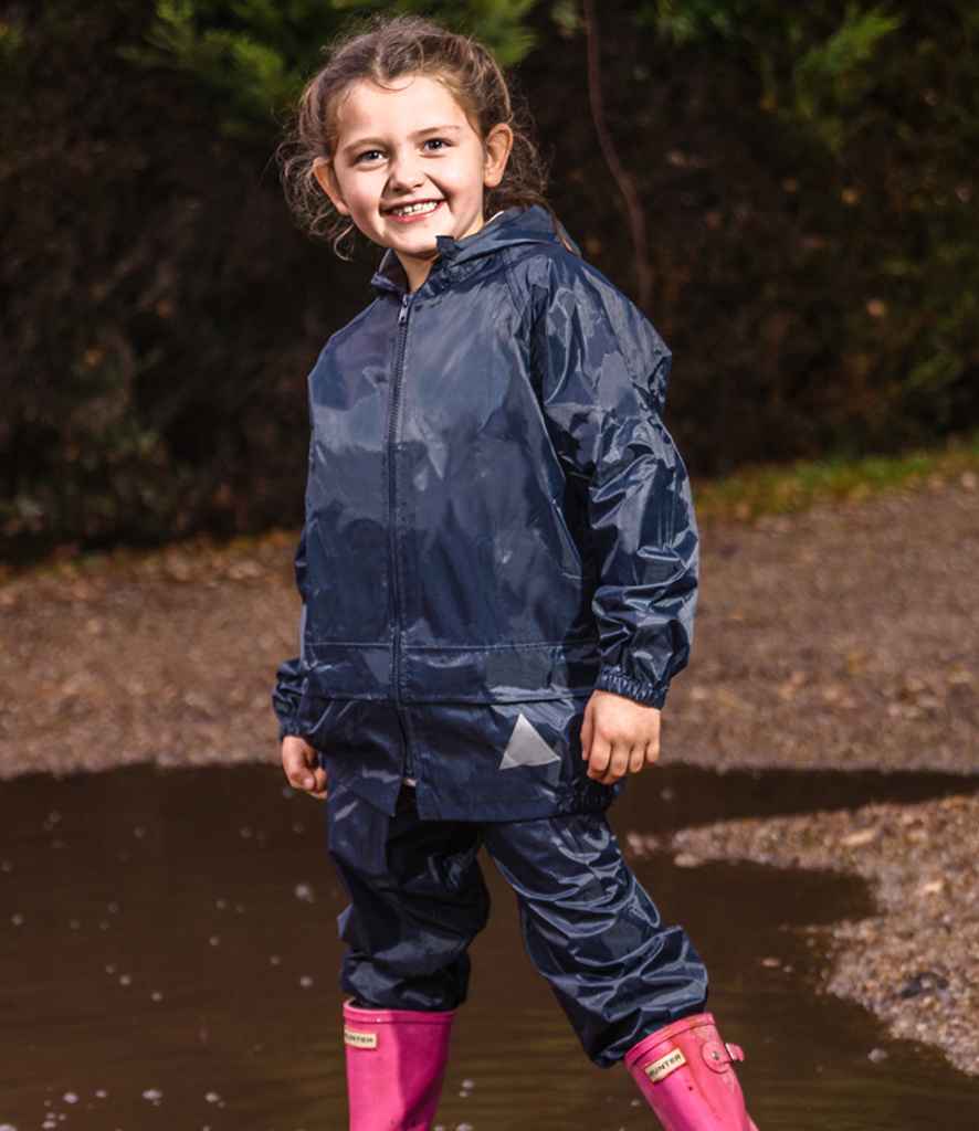 A young child stands outdoors on wet ground wearing the Result Kids Waterproof Jacket/Trouser Suit in navy blue by Result, paired with bright pink rain boots. Trees and greenery appear in the background.