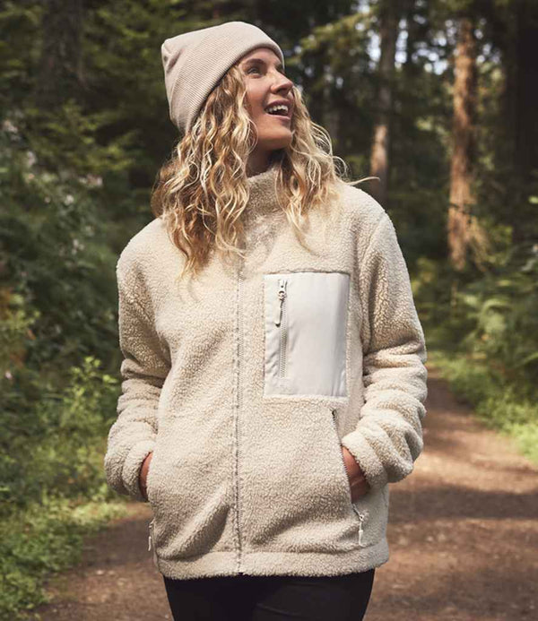 Wearing the Ravine Unisex Full Zip Borg Fleece Jacket, a person in a beige beanie stands on a forest path with hands in their pockets, looking up and smiling amidst lush green trees and plants.