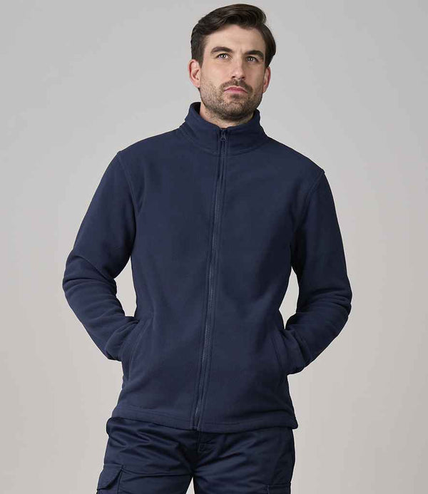 A man with short hair and a beard stands against a plain background, hands in the pockets of a Pro RTX Pro Fleece Jacket by Pro RTX—a dark blue unisex full-zip jacket featuring an adjustable hem. He looks slightly to the side.