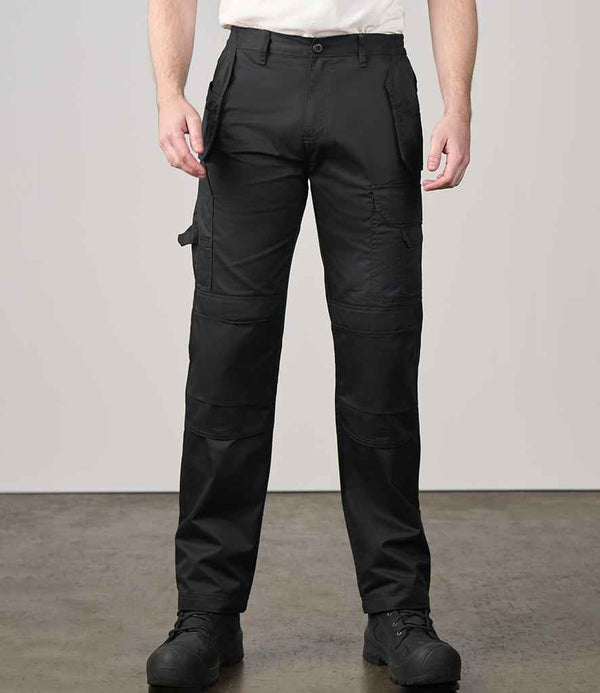 A person wearing Pro RTX Pro Tradesman Trousers by Pro RTX with tradesman pockets and black shoes stands indoors on a polished concrete floor against a plain light-colored wall, shown from shoulders to feet.