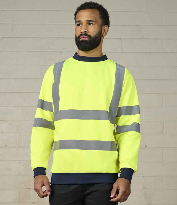 A man with a beard stands against a light brick wall, wearing the Pro RTX High Visibility Two Tone Sweatshirt by Pro RTX High Visibility, featuring reflective bands, neon yellow fabric, and dark cuffs and collar.