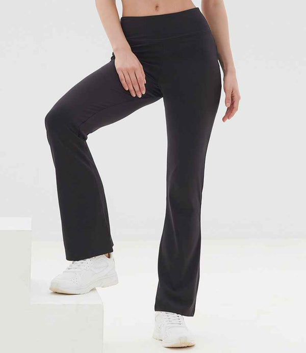 A person models SF Clothing Ladies Flared Leggings—black, high-waisted, soft-touch flared pants—from SF Clothing with white sneakers, posing with one leg bent and the other straight against a light background; upper body is partially out of frame.