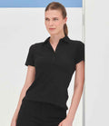 A woman with light skin and brown hair tied back models the SF Clothing Ladies Stretch Piqué Polo Shirt— a slim fit, black short-sleeve polo with a self-fabric collar and shaped side seams, against a minimal white background accented in blue.