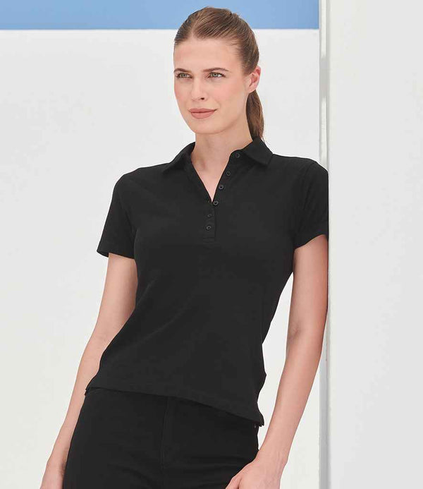 A woman with light skin and brown hair tied back models the SF Clothing Ladies Stretch Piqué Polo Shirt— a slim fit, black short-sleeve polo with a self-fabric collar and shaped side seams, against a minimal white background accented in blue.