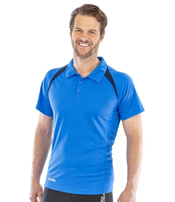 A man with short brown hair and a beard stands smiling in a Spiro Team Spirit Polo Shirt by Spiro. The blue, quick-drying shirt has black shoulder accents and is paired with black pants against a white background.