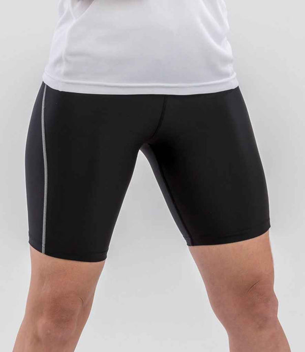 A person wears Spiro Ladies Bodyfit Base Layer Shorts by Spiro—black with a vertical gray stripe on the left, made from lightweight breathable quick-dry fabric—standing against a plain light background.