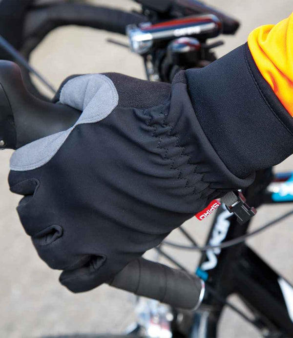 A person wearing Spiro Long Winter Gloves by Spiro—black and gray gloves with polyurethane palm patches—grips a bicycle handlebar; the orange sleeve, brake lever, and part of the bike frame are visible in the background.