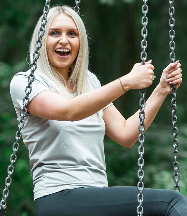 A woman with long blonde hair sits on a swing, smiling and holding the chains. She wears a light gray Spiro Impact Ladies Softex® T-Shirt by Spiro and dark pants. The background features blurred greenery.