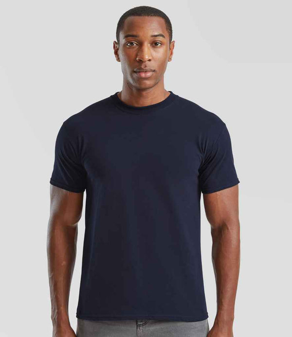 A man stands against a plain light gray background, wearing the Fruit of the Loom Super Premium T-Shirt in navy blue with a tubular body and Cotton/Lycra collar, paired with gray pants. He faces forward with a neutral expression.