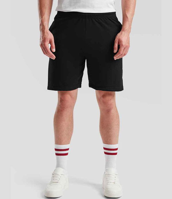 A person wears a white t-shirt, Fruit of the Loom Lightweight Shorts in black with an elasticated waistband and side pockets, white socks with red stripes, and white sneakers, standing against a plain light gray background.