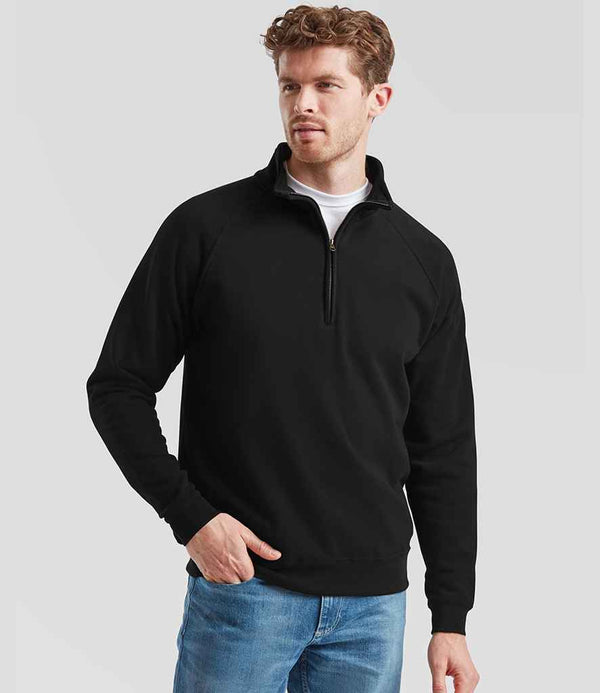 A man with short, curly brown hair and a trimmed beard stands against a plain background, wearing light blue jeans and the Fruit of the Loom Classic Zip Neck Sweatshirt by Fruit of the Loom. One hand is in his pocket.