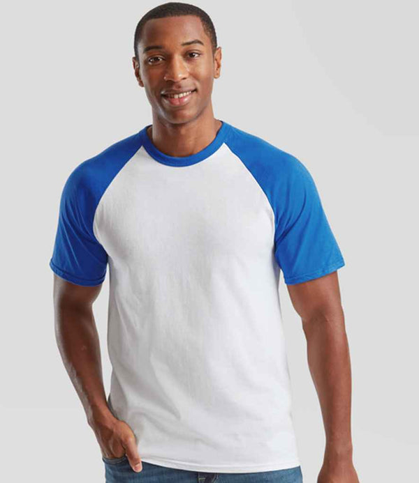 A man stands against a light gray background, smiling as he wears the Fruit of the Loom Contrast Baseball T-Shirt with contrast raglan sleeves and a ribbed collar, paired with blue jeans. One hand is by his side while the other touches his jeans.