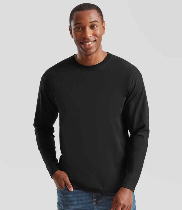 A man stands against a light gray background, smiling at the camera with one hand in his pocket. He wears blue jeans and a Fruit of the Loom Original Long Sleeve T-Shirt by Fruit of the Loom, featuring a ribbed collar.