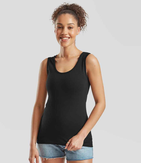 A woman with curly hair smiles at the camera, wearing the Fruit of the Loom Lady Fit Value Vest—a black sleeveless tank top with shaped side seams—paired with denim shorts against a light gray background.