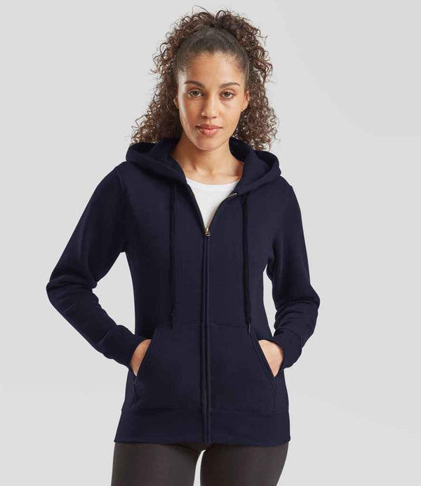 A person with curly hair wears a Fruit of the Loom Premium Lady Fit Zip Hooded Jacket in navy blue, featuring ribbed cuffs and front pockets, paired with black pants, standing against a plain light gray background.