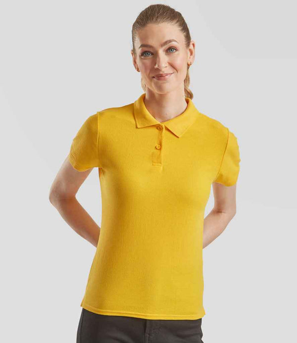 A woman stands against a plain light gray background, wearing a Fruit of the Loom Lady Fit Piqué Polo Shirt in yellow with cuffed sleeves and black pants, her hands placed behind her back.