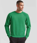 A man faces slightly sideways in a light gray setting, wearing the Fruit of the Loom Classic Raglan Sweatshirt in plain green with a ribbed collar and black pants.