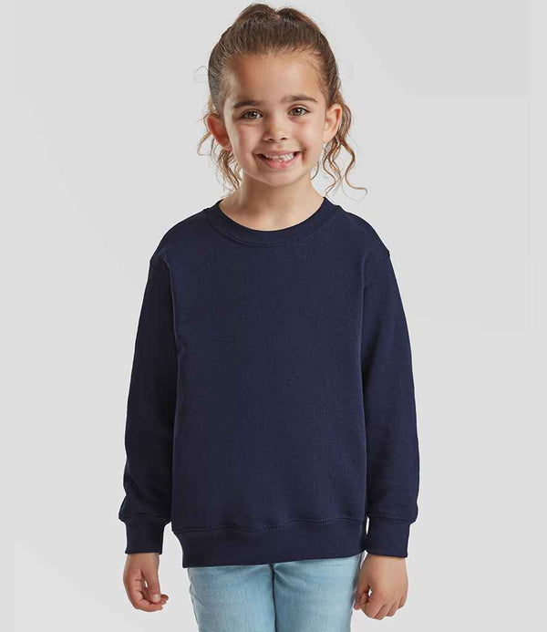 A young girl with curly hair smiles at the camera, wearing a navy blue Fruit of the Loom Kids Classic Drop Shoulder Sweatshirt and light blue jeans, standing against a plain light gray background.