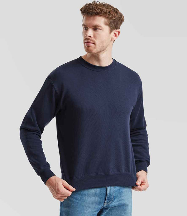 A man wearing the Fruit of the Loom Classic Drop Shoulder Sweatshirt in navy and light blue jeans stands against a light gray background, looking off to the side.