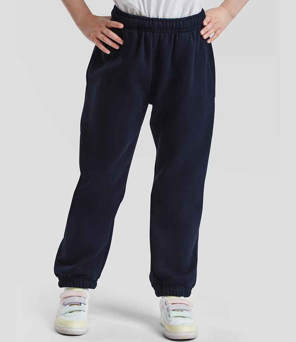 A child stands with hands on hips, wearing Fruit of the Loom Kids Premium Jog Pants in navy blue featuring an elastic waistband, cuffs, and side pockets. The look is completed with white sneakers and a plain light gray background.