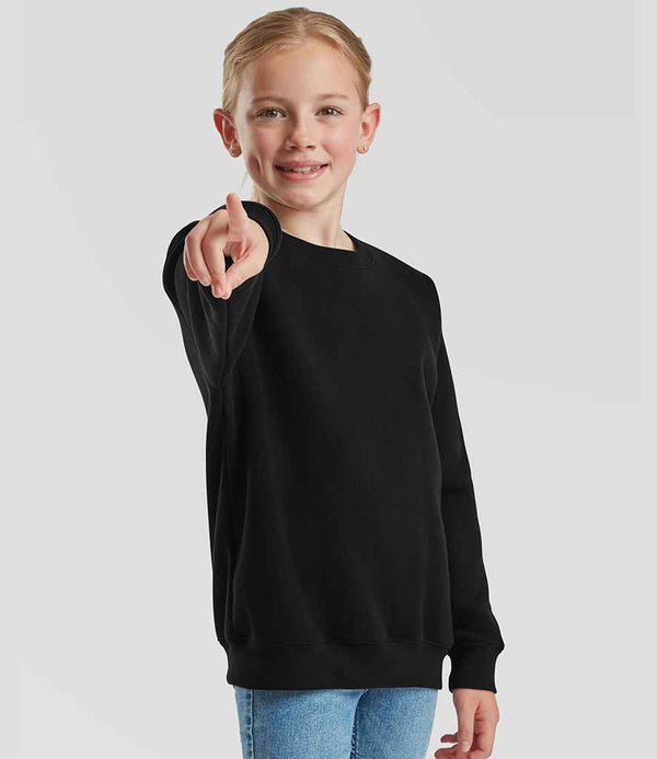 A smiling young person with light hair stands against a plain background, wearing blue jeans and a black Fruit of the Loom Kids Premium Drop Shoulder Sweatshirt, featuring twin needle stitching, while pointing forward.
