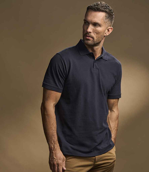A man with short hair and a trimmed beard wears the Tee Jays Heavy Cotton Piqué Polo Shirt by Tee Jays in navy blue, paired with tan pants. He stands against a plain brown background, looking slightly to his left.