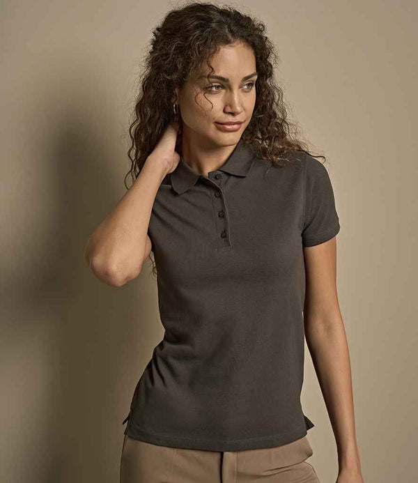 A woman with curly hair wears a fitted Tee Jays Ladies Heavy Cotton Piqué Polo Shirt with short sleeves, four-button placket, and dark color, paired with light pants. She stands against a beige background, looking sideways and touching her neck.