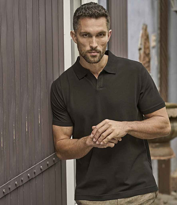 A man with short hair and facial hair stands before a wooden door, wearing the Tee Jays Luxury Stretch V Neck Polo Shirt by Tee Jays in black, tailored fit with a flat knit collar, paired with light-colored pants, hands loosely clasped.
