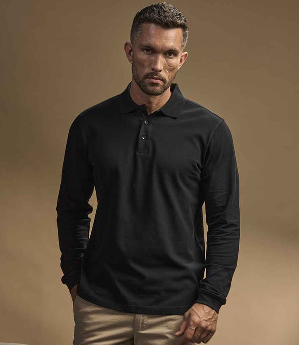 A man with short hair and a beard wears the Tee Jays Luxury Stretch Long Sleeve Polo Shirt in black and beige pants. He stands against a plain brown background, facing the camera with a neutral expression.
