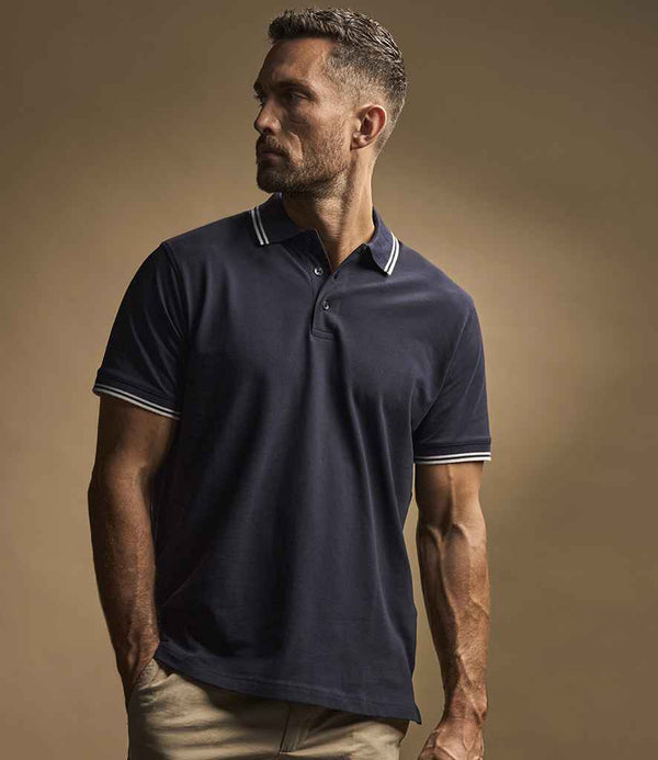 A man with short hair and a trimmed beard wears the Tee Jays Luxury Stretch Tipped Polo Shirt in navy by Tee Jays, paired with beige pants, standing against a neutral background and looking to the side.