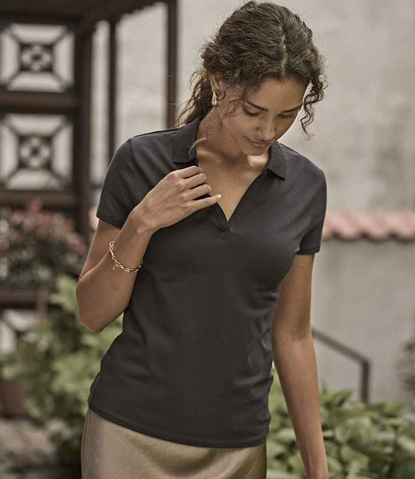 A woman with curly hair wears the Tee Jays Ladies Luxury Stretch V Neck Polo Shirt, a short-sleeved black top with a flat knit collar and tailored fit. She stands outdoors, gold bracelet on her wrist, surrounded by plants and buildings.