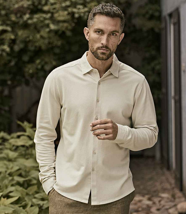 A man with short dark hair and a trimmed beard stands outdoors in a Tee Jays Luxury Organic Stretch Long Sleeve Shirt by Tee Jays, light beige in color, paired with brown pants. He looks at the camera with one hand raised, plants in the background.