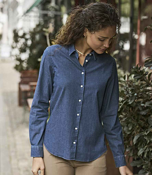 A woman with curly hair stands outdoors, surrounded by plants and seating, wearing a Tee Jays Ladies Denim Shirt in garment washed blue with modern cuffs, paired with light brown pants.