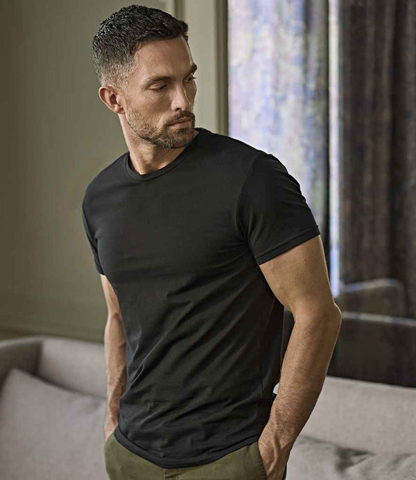 A man with short dark hair and a trimmed beard stands indoors, hands in his pockets, wearing a Tee Jays Stretch T-Shirt in black and green pants, looking to the side.