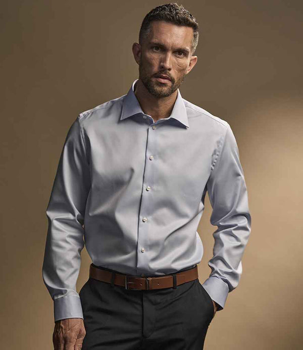 A man with short hair and a trimmed beard is wearing the Tee Jays Luxury Comfort Fit Long Sleeve Oxford Shirt in light blue by Tee Jays, paired with black pants and a brown belt, standing against a plain, neutral background.