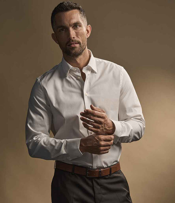 A man with short hair and a beard wears a white Tee Jays Luxury Stretch Long Sleeve Shirt with adjustable cuffs, tucked into dark trousers with a brown belt, facing the camera and adjusting his sleeve against a warm brown background.