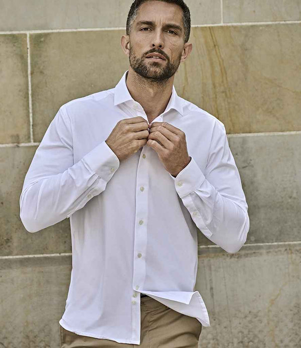 A man with short hair and a beard buttons a white Tee Jays Active Stretch Long Sleeve Shirt, known for its moisture-wicking fabric. He stands against a stone wall in beige pants, looking at the camera with a neutral expression.
