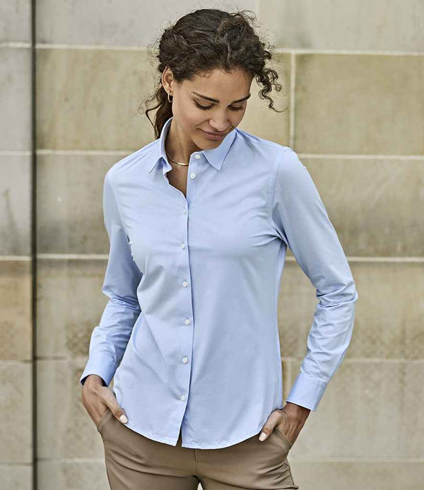 A woman wearing the Tee Jays Ladies Active Long Sleeve Stretch Shirt by Tee Jays in light blue and tan pants stands against a beige stone wall, hands in her pockets, looking down with a slight smile.