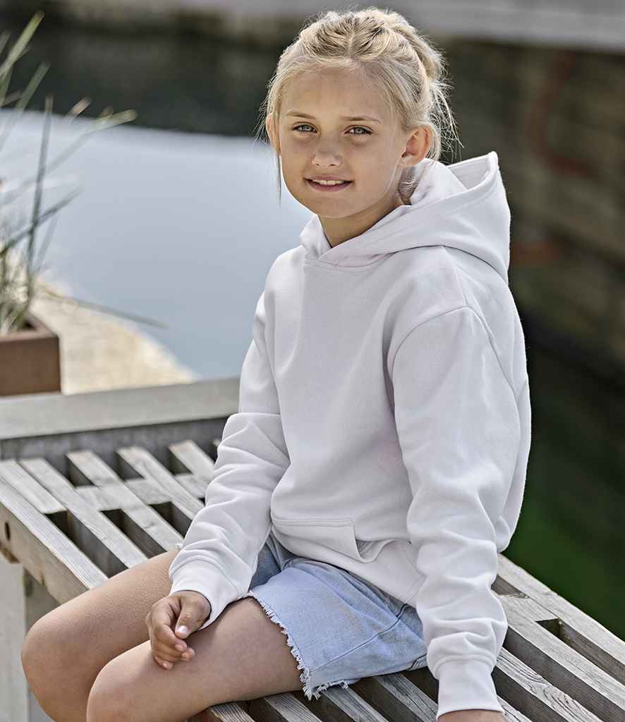 A young girl with blonde hair in a ponytail sits on a wooden bench by the water, wearing a Tee Jays Kids Unlabeled Hooded Sweatshirt and light blue denim shorts. She looks at the camera with a slight smile.