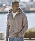 A man wears a Tee Jays Unlabeled Hooded Full Zip Sweatshirt in light brown over a white t-shirt, standing outdoors with hands in pockets and looking left. The background is blurred outdoor scenery.