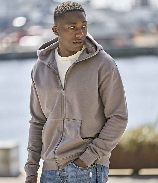 A man wears a Tee Jays Unlabeled Hooded Full Zip Sweatshirt in light brown over a white t-shirt, standing outdoors with hands in pockets and looking left. The background is blurred outdoor scenery.
